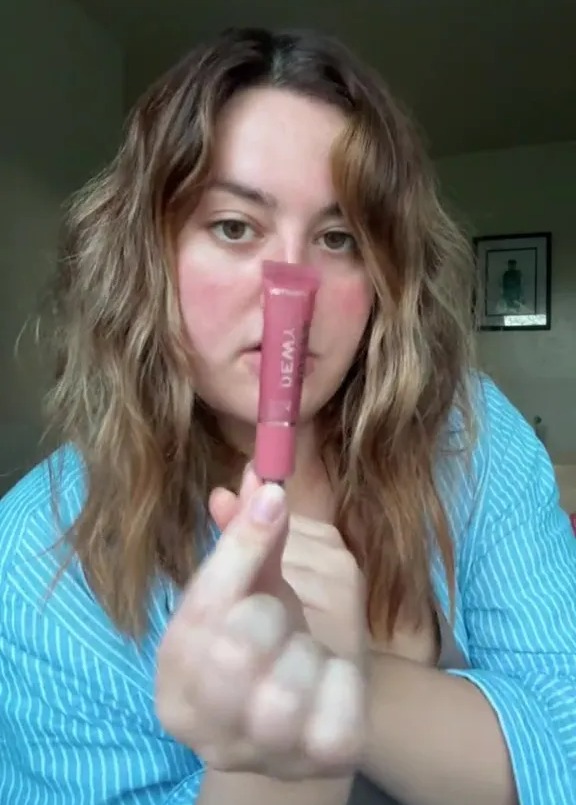 a woman in a blue shirt is holding a tube of lip gloss