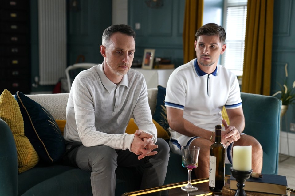 two men sit on a couch with a bottle of wine on the table