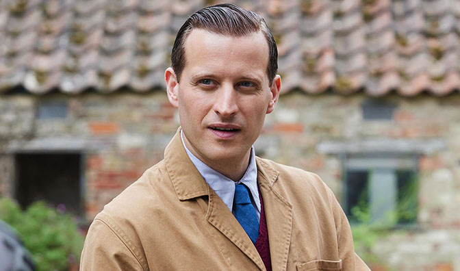 a man in a tan jacket and blue tie stands in front of a brick building