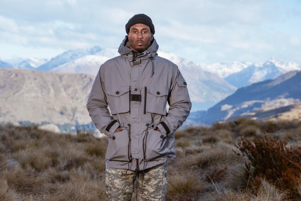 a man in a grey jacket stands in a field with mountains in the background