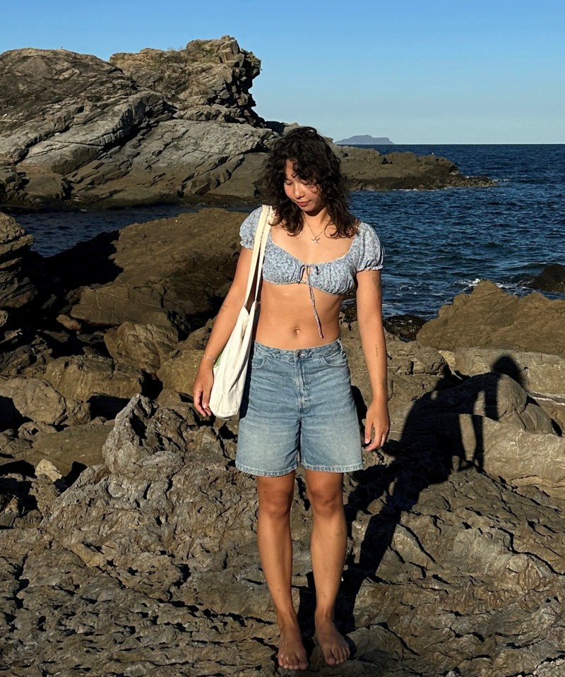 a woman in a bikini top and shorts stands on a rocky beach