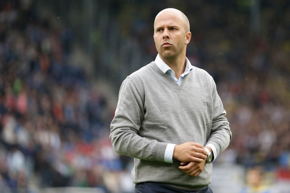 a bald man in a grey sweater stands on a soccer field