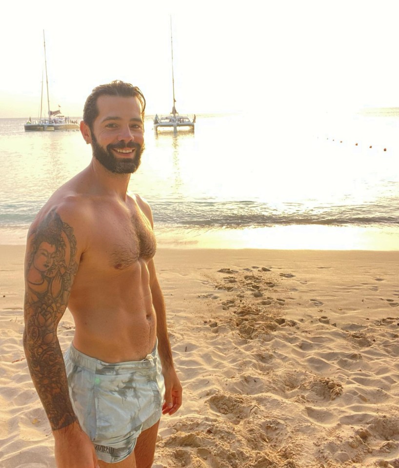 a shirtless man stands on a beach with a boat in the background