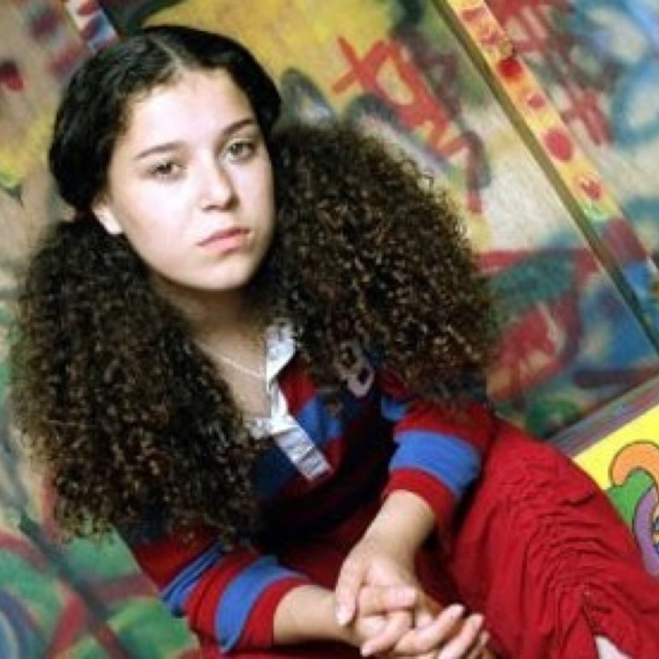 a young girl with curly hair is sitting in front of a wall with graffiti on it .