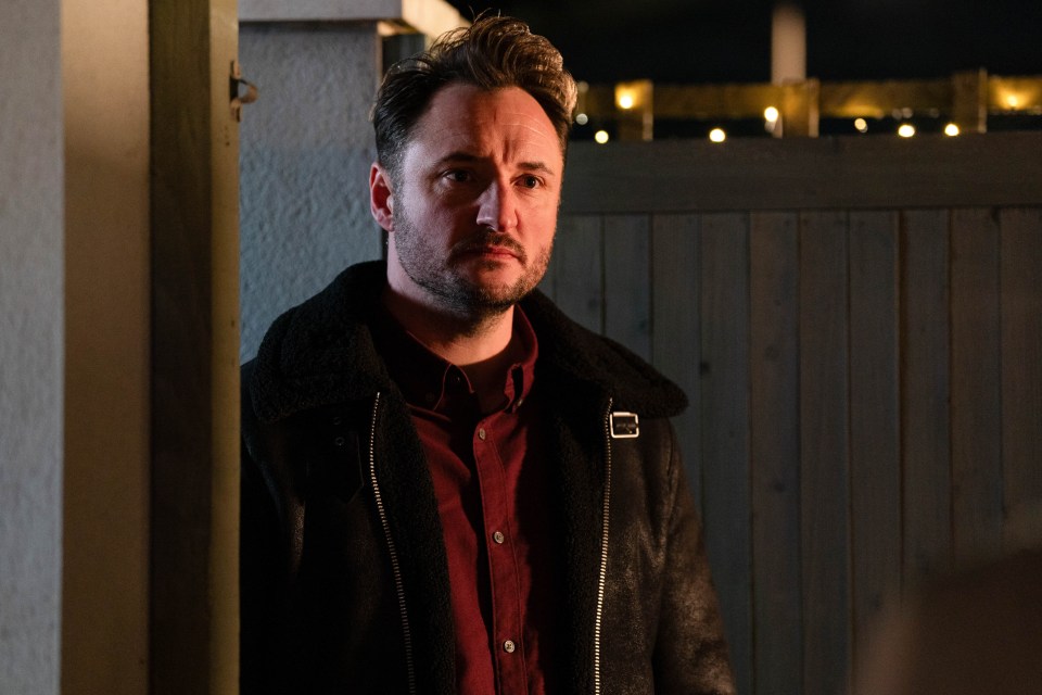 a man in a black jacket stands in front of a wooden fence