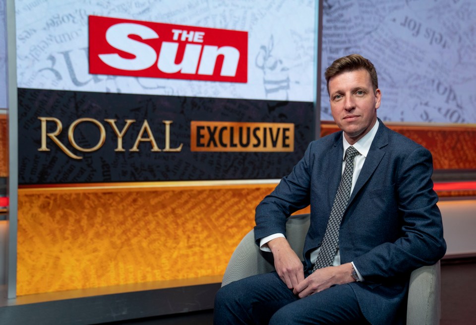 a man in a suit sits in front of a sign for the sun