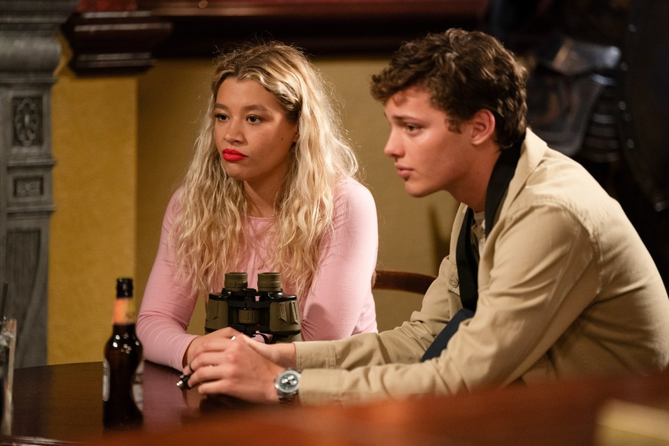 a man and a woman sit at a table with binoculars and a bottle of beer