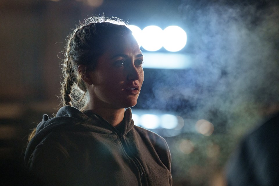 a woman in a hoodie stands in a dark room with smoke coming out of her mouth