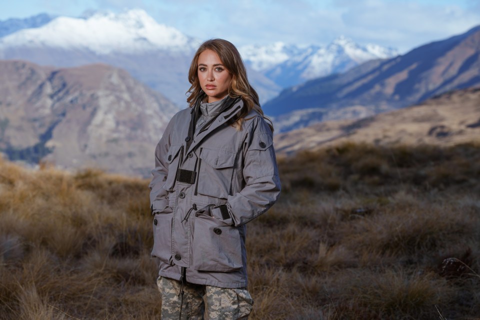 a woman in a military uniform stands in a field with mountains in the background