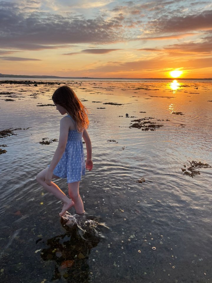 a little girl in a blue dress stands in the water at sunset