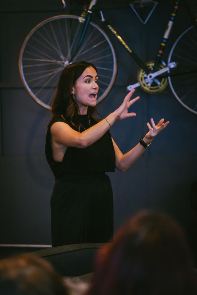 a woman in a black dress stands in front of a wall with a bicycle in the background