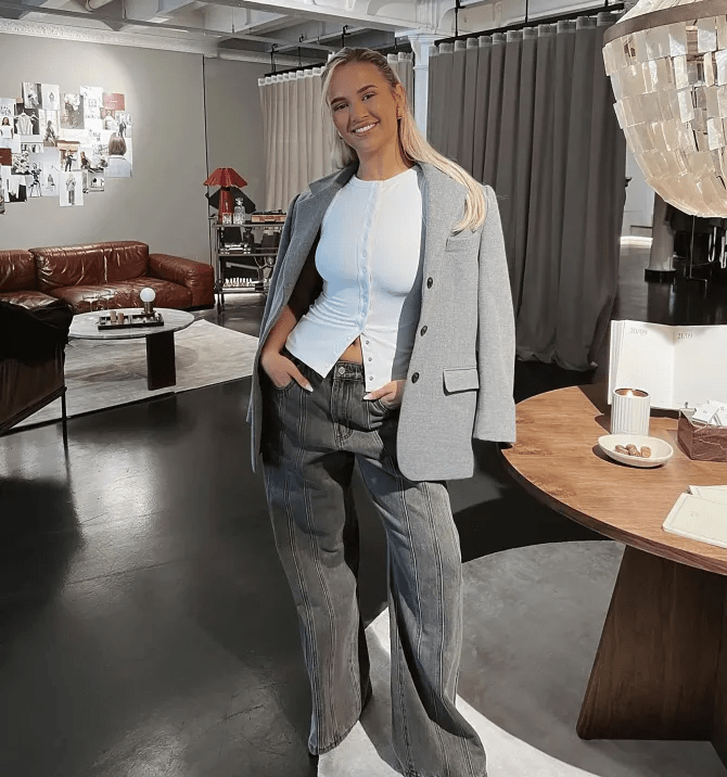 a woman wearing a grey jacket and jeans stands in a living room