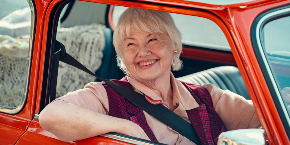 an elderly woman wearing a seat belt smiles while sitting in a red car