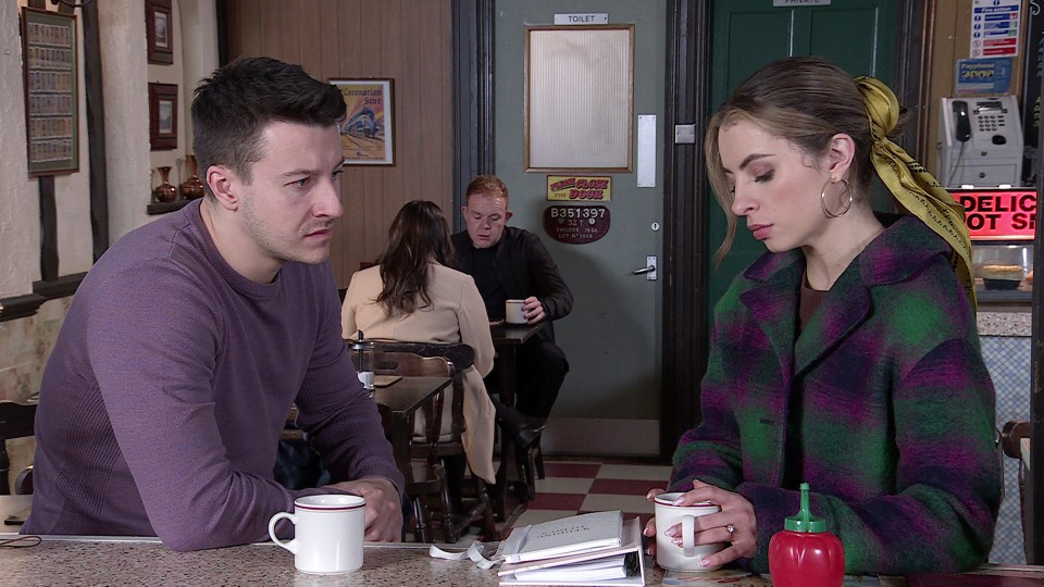 a man and a woman sit at a table in front of a sign that says deli hot sandwiches