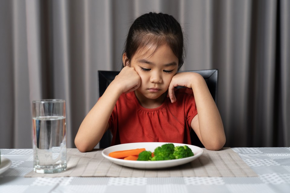 a little girl sits at a table with a plate of vegetables and a glass of water