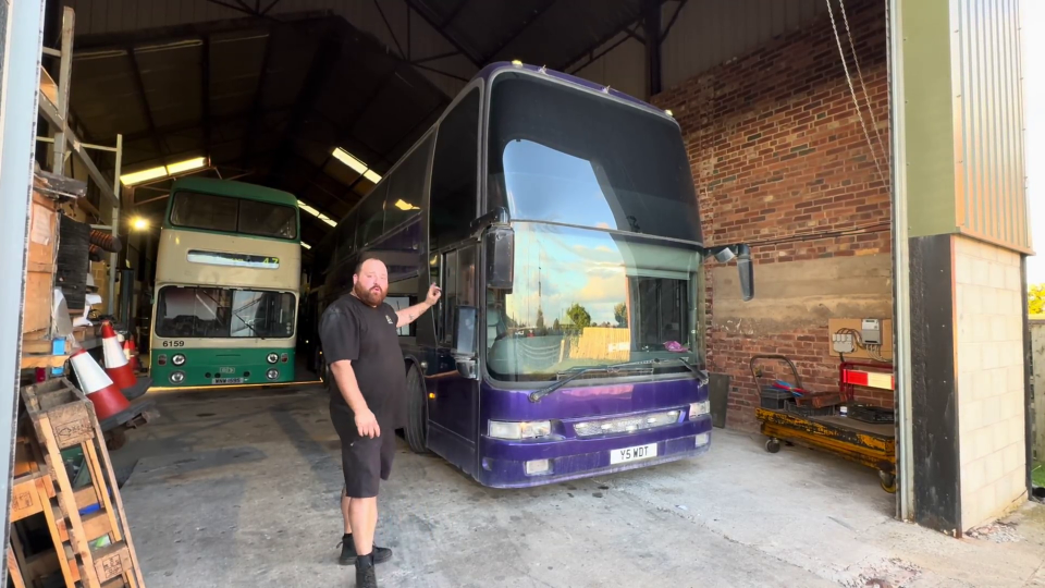 a man stands in front of a purple double decker bus with a license plate that says n15 k01