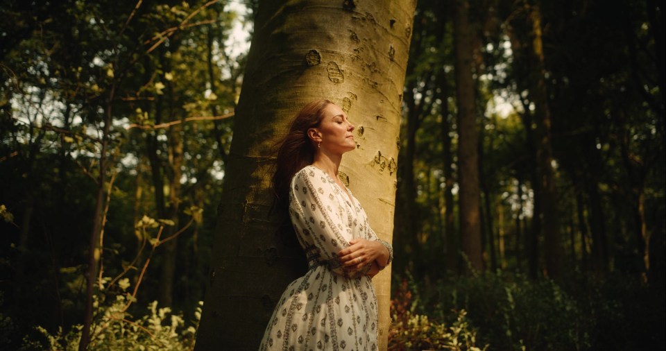 a woman in a white dress leans against a tree in the woods