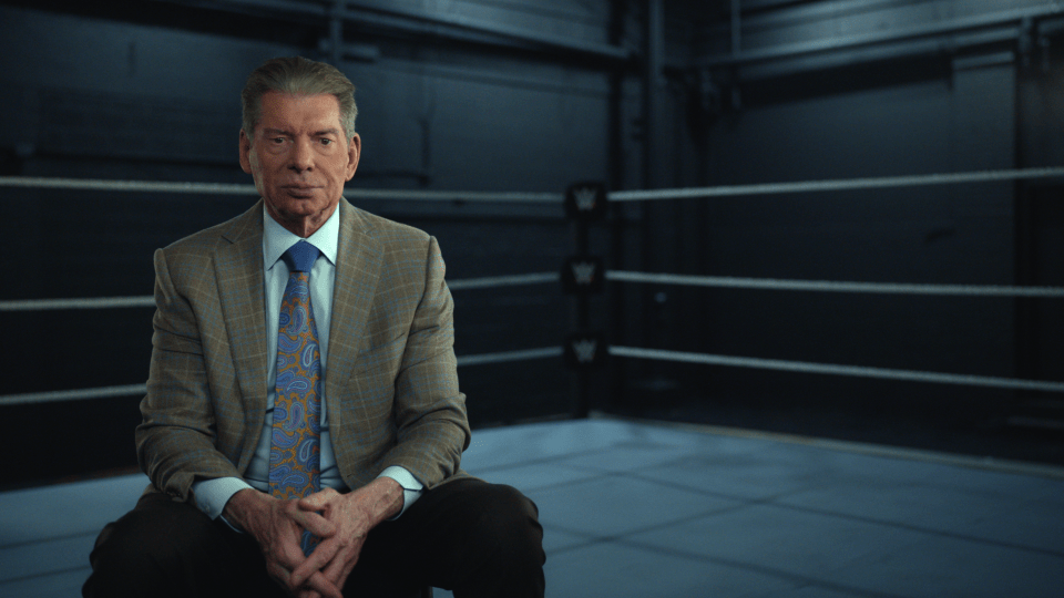 a man in a suit and tie sits in front of a wrestling ring