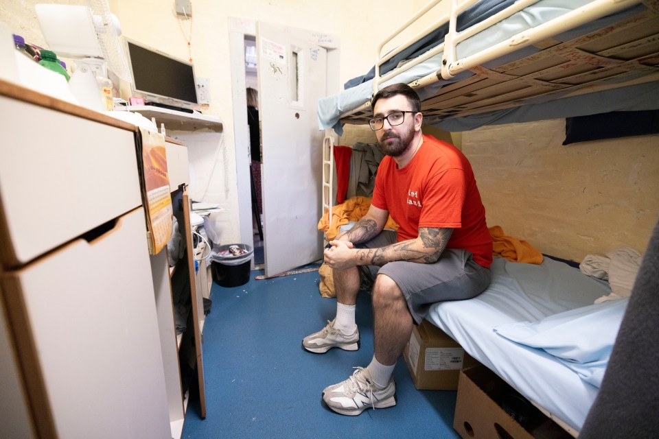 a man sits on a bunk bed in a room with a red shirt that says ' prison ' on it