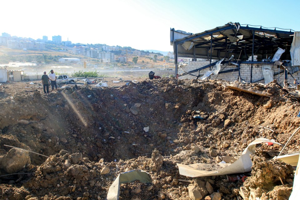 People stand by an impact crater next to a destroyed warehouse - believed to belong to Hezbollah - at the site of an Israeli air strike in Jiyeh