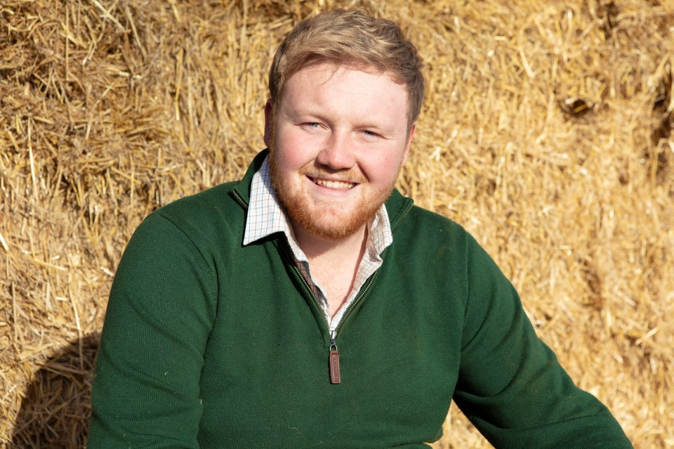 a man in a green sweater stands in front of a pile of hay