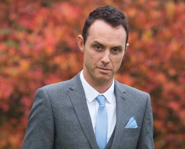 a man in a suit and tie stands in front of a bush with red leaves