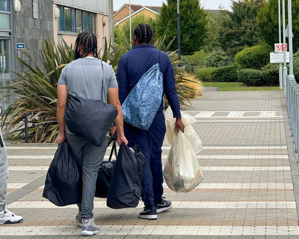 two men carrying bags walk down a sidewalk in front of a no parking sign