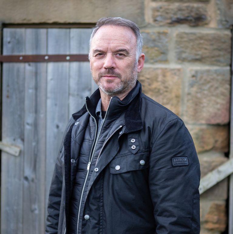 a man wearing a black barbour jacket stands in front of a stone wall