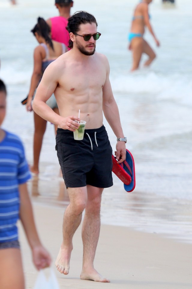 a shirtless man walking on the beach holding a drink and a pair of flip flops