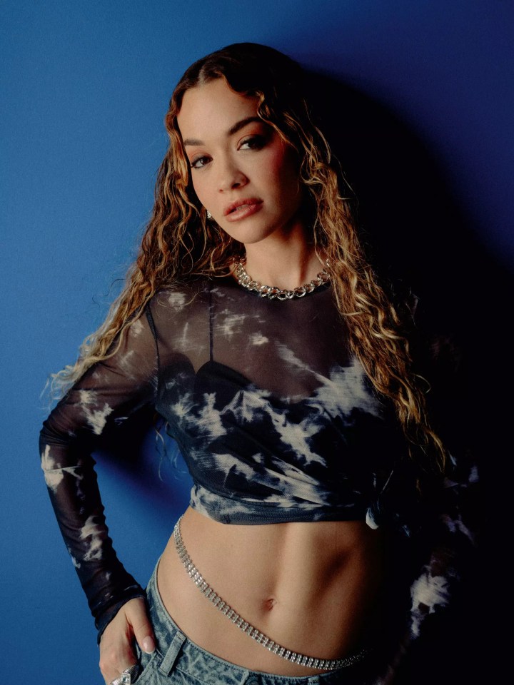 a woman wearing a tie dye top and jeans stands in front of a blue wall