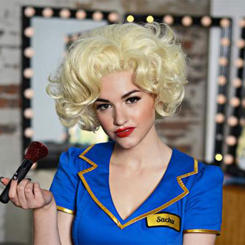 a woman in a blonde wig is holding a makeup brush in front of a mirror .