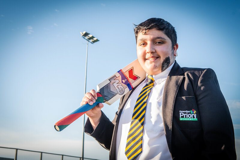 a boy in a suit and tie is holding a cricket bat with the word priority on his jacket