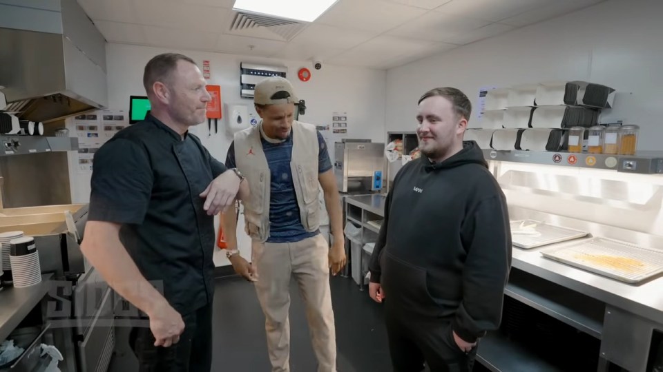 three men are standing in a kitchen and one of them is wearing a black hoodie that says ' hoodie ' on it