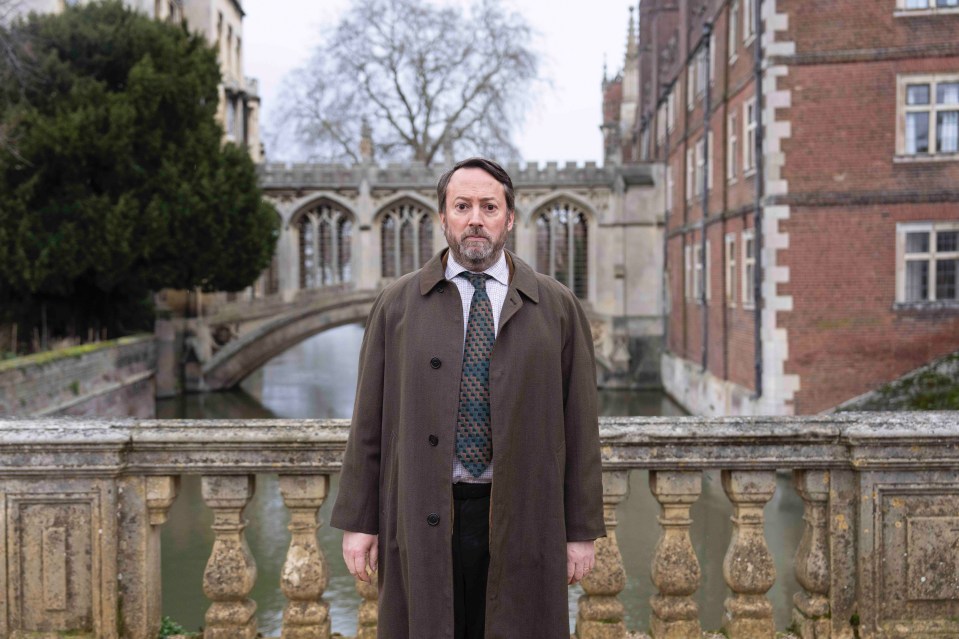 a man in a trench coat and tie stands on a bridge over a river