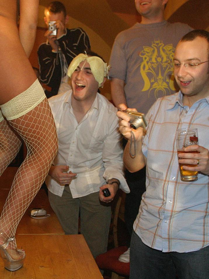 One tourist wears a bra on his head while a woman in fishnet stockings and platforms stands on a table as another man holds a beer and a camera