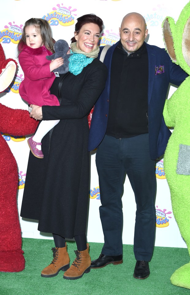 a woman holding a little girl stands next to a man and a green stuffed animal