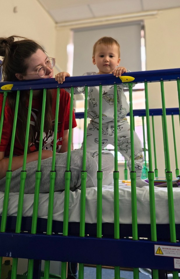 a woman in a red christmas shirt sits next to a baby in a crib