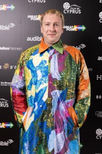 a man in a colorful coat stands in front of a wall that says film in cyprus