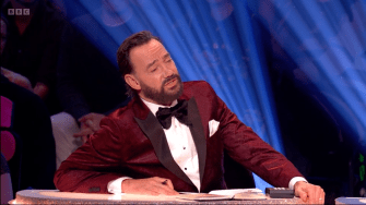 a man in a red tuxedo sits at a table with bbc written on the bottom