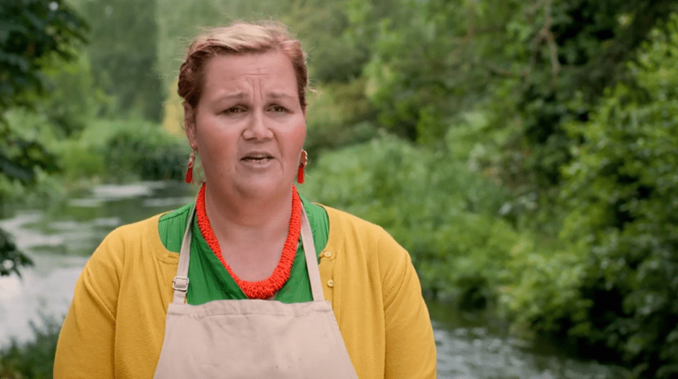 a woman wearing a yellow cardigan and an apron stands in front of a river