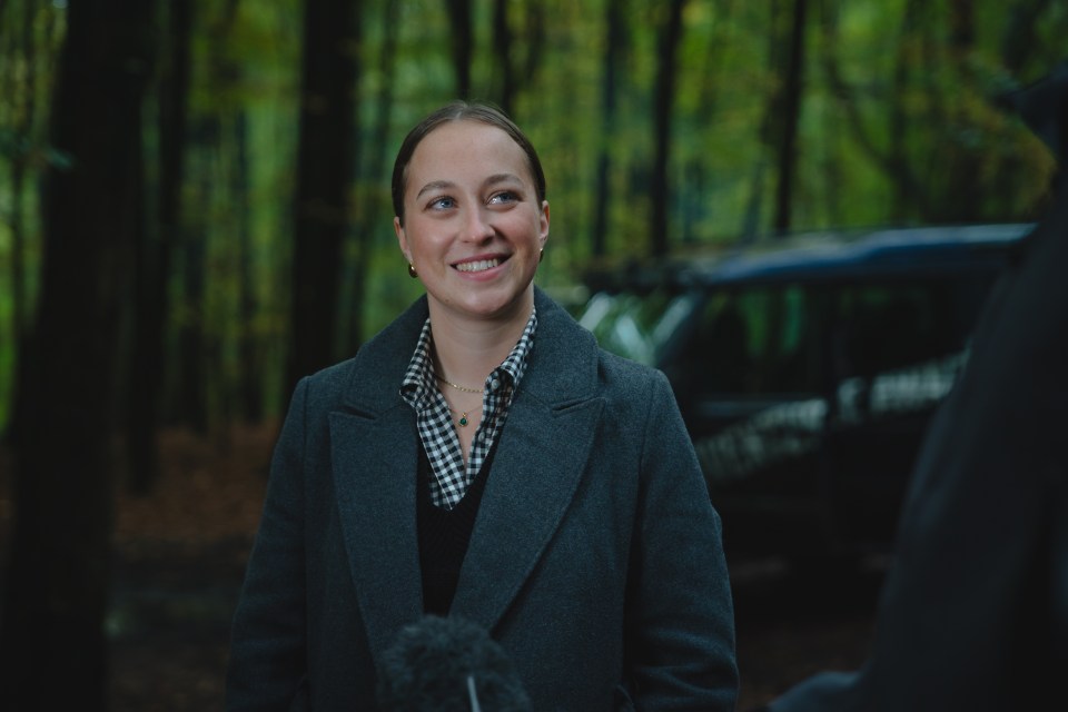 a woman in a grey coat smiles in front of a car