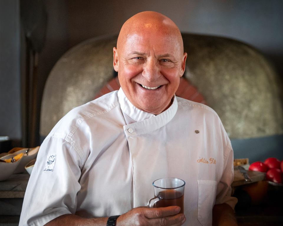 a smiling bald man wearing a white chef 's jacket with the name alla on it