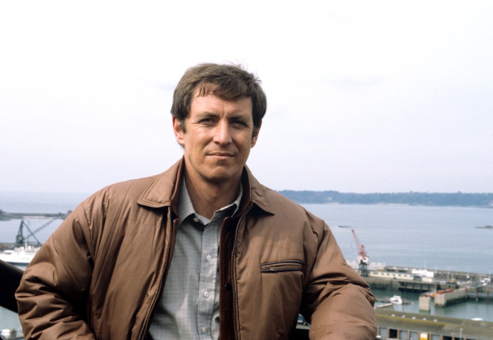 a man in a brown jacket stands in front of a body of water