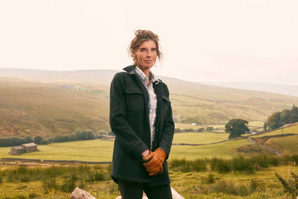 a woman in a black jacket and brown gloves stands in a field