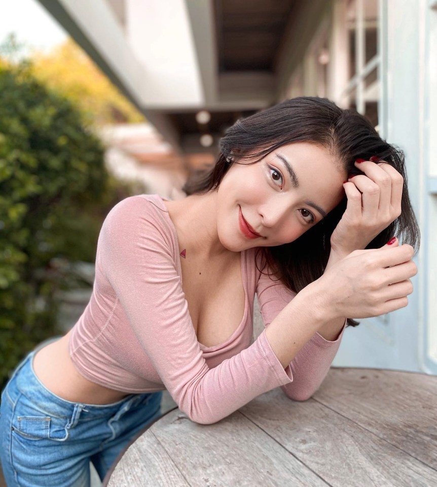 a woman in a pink top and blue jeans leans on a wooden table