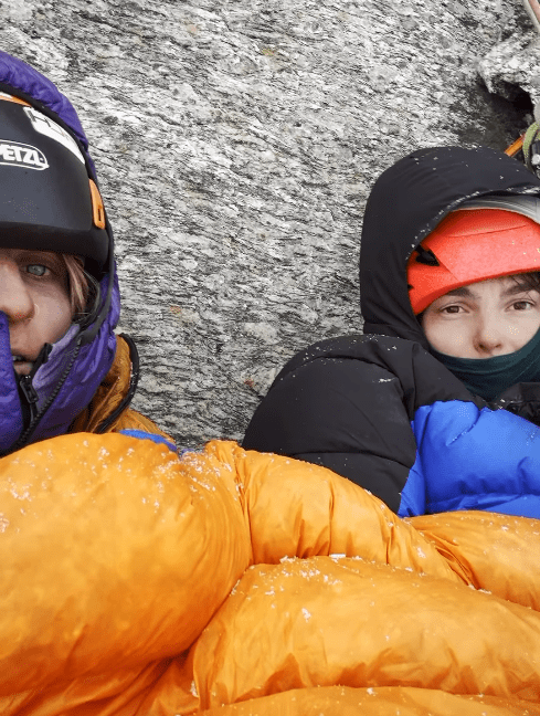 a person wearing a petzl helmet sits next to another person in a sleeping bag