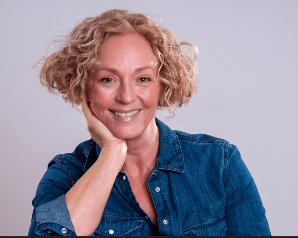 a woman in a blue denim shirt smiles with her hand on her chin