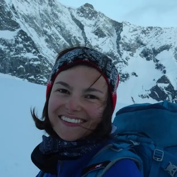 a woman wearing a headband and a blue backpack smiles in front of a snowy mountain