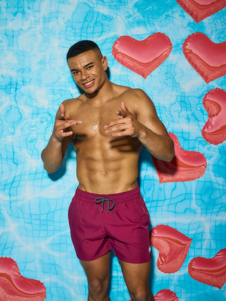 a shirtless man in purple swim trunks stands in front of pink heart shaped balloons