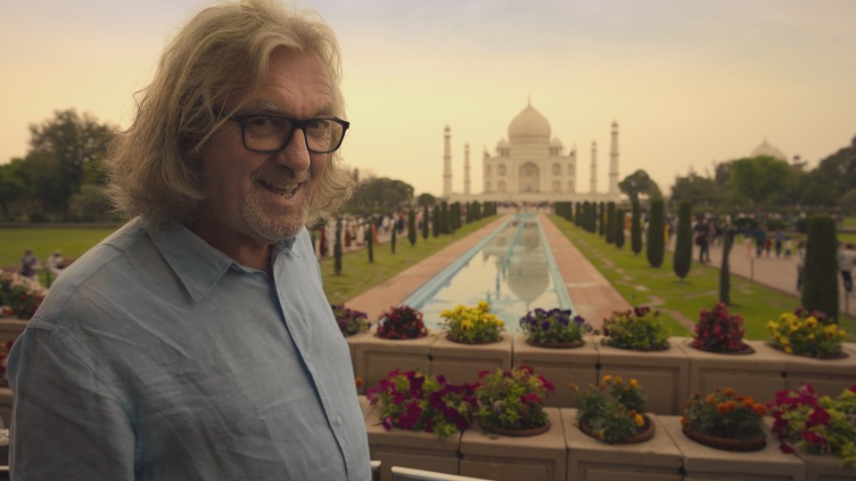 a man with long hair and glasses stands in front of a large building